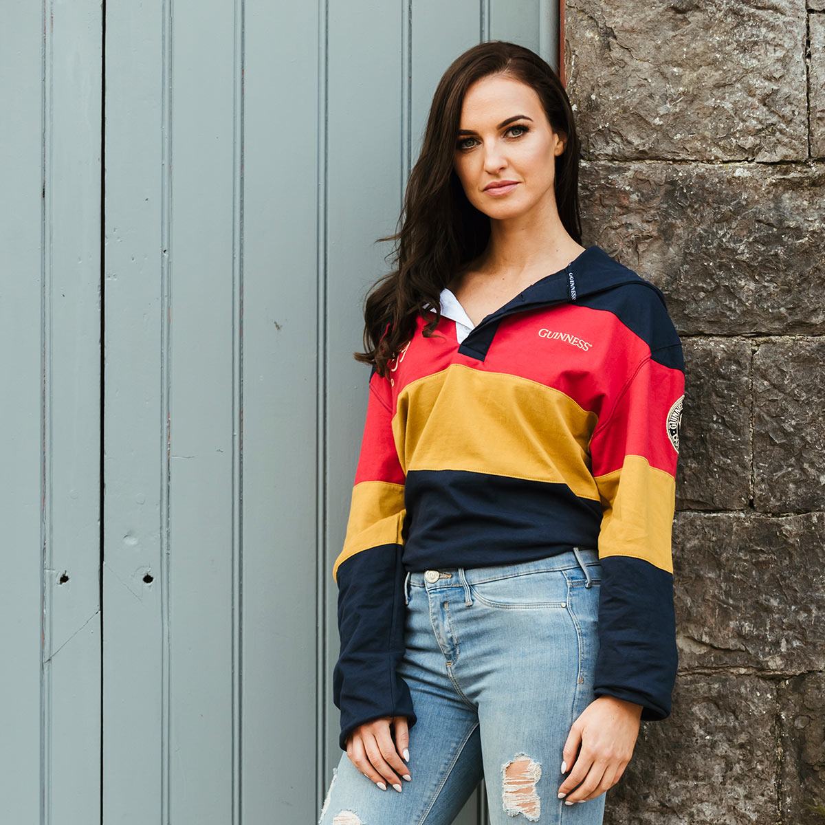 A woman in ripped jeans leaning against a wall, wearing a Guinness Hooded Rugby Jersey.