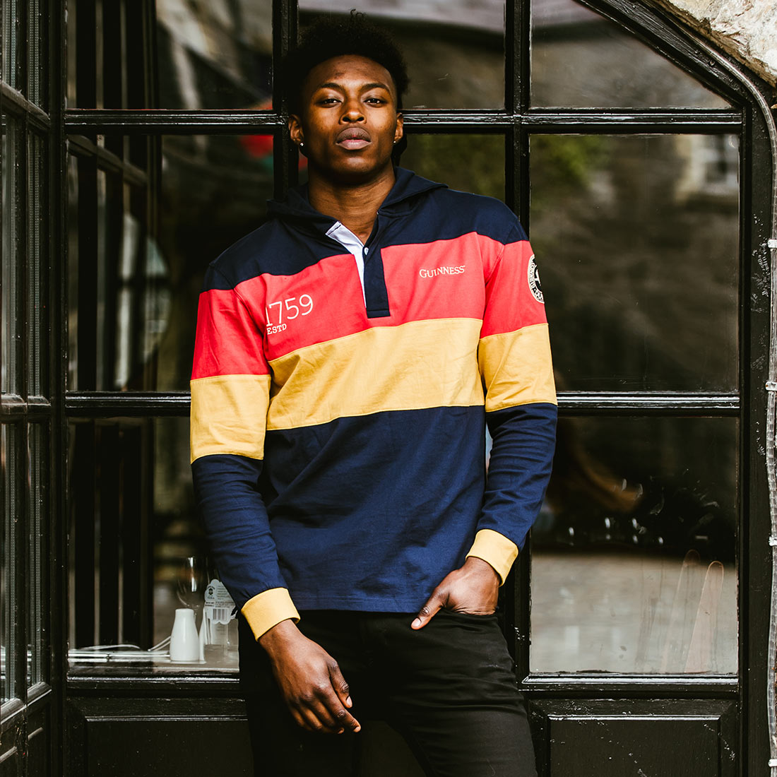 A man wearing a colorful hoodie, reminiscent of a Guinness Hooded Rugby Jersey, standing in front of a window.