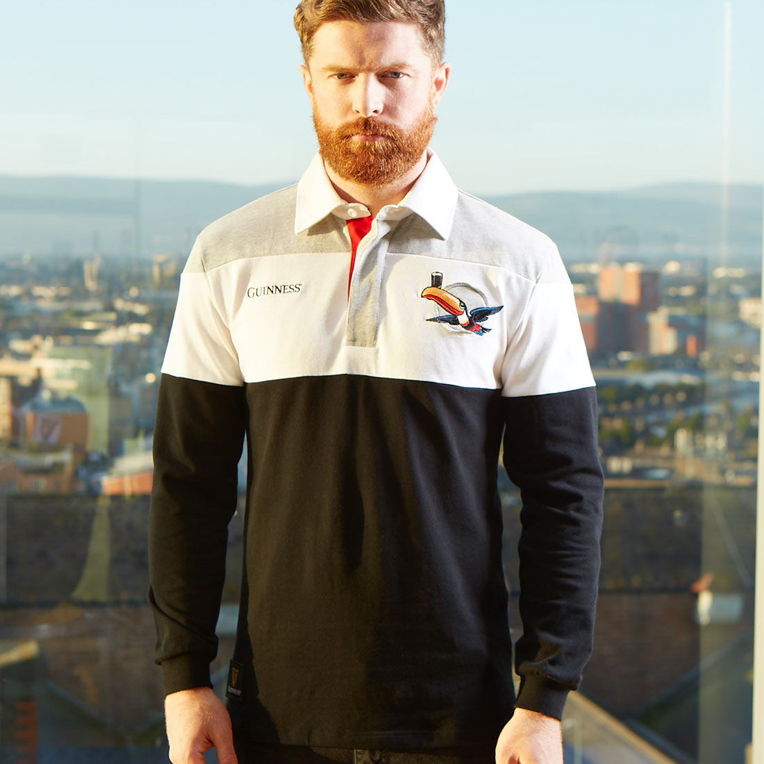 A bearded man in a Guinness Toucan Rugby Jersey standing in front of a city, showcasing branding for Guinness UK.