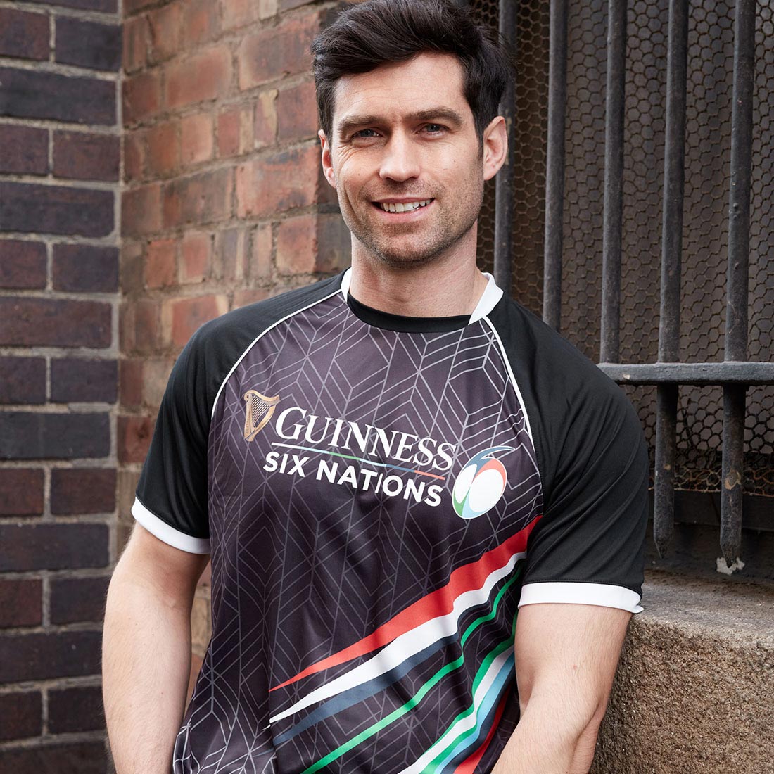 A rugby fan wearing a Guinness Six Nations Performance T-Shirt leaning against a brick wall.