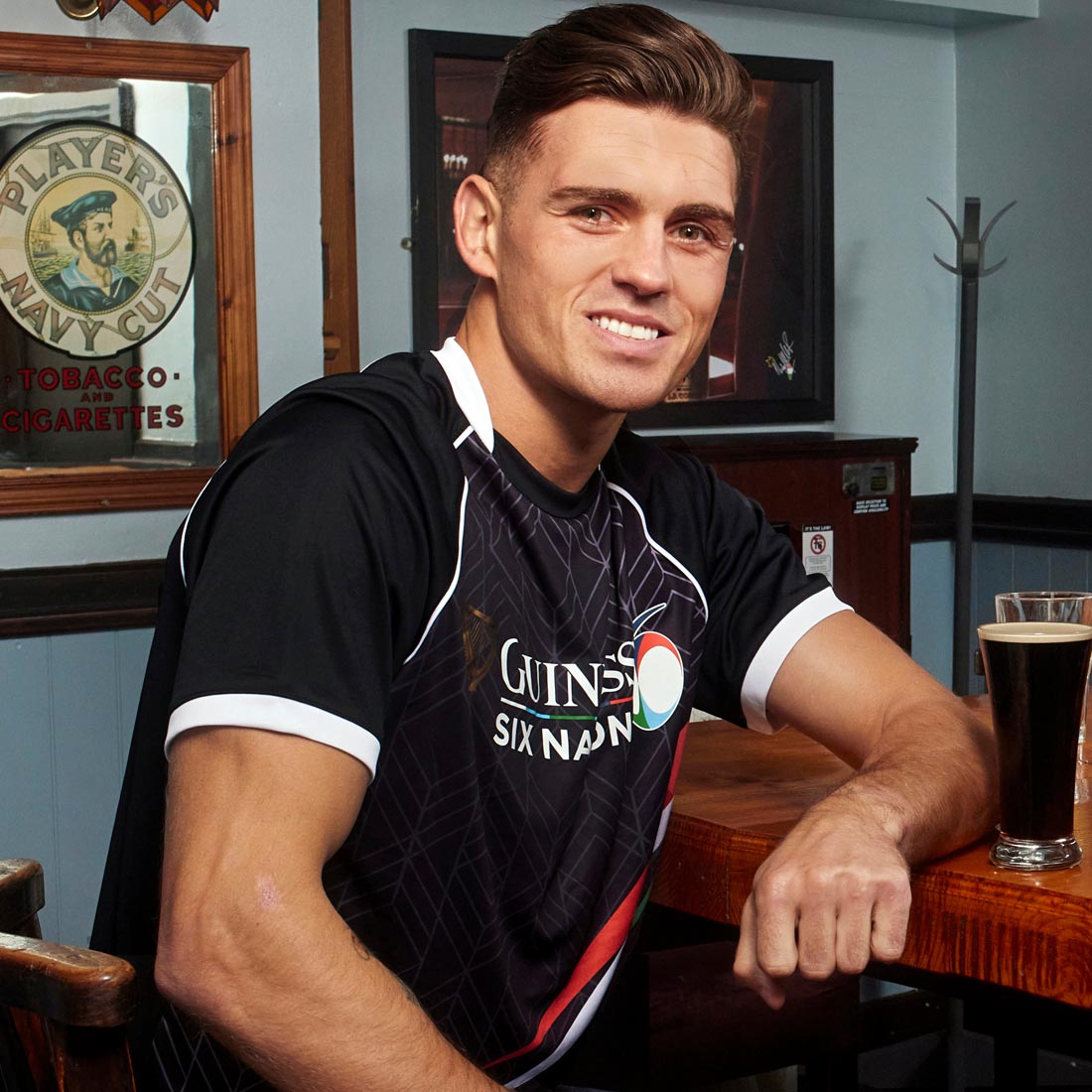 A rugby fan, wearing a Guinness 6 Nations Performance T-Shirt, is sitting at a table with a beer.