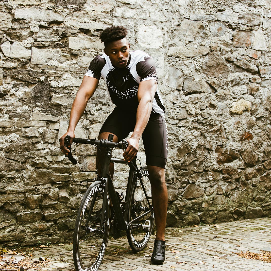 A man wearing a Guinness UK Cycling Jersey is riding a bike in front of a stone wall.