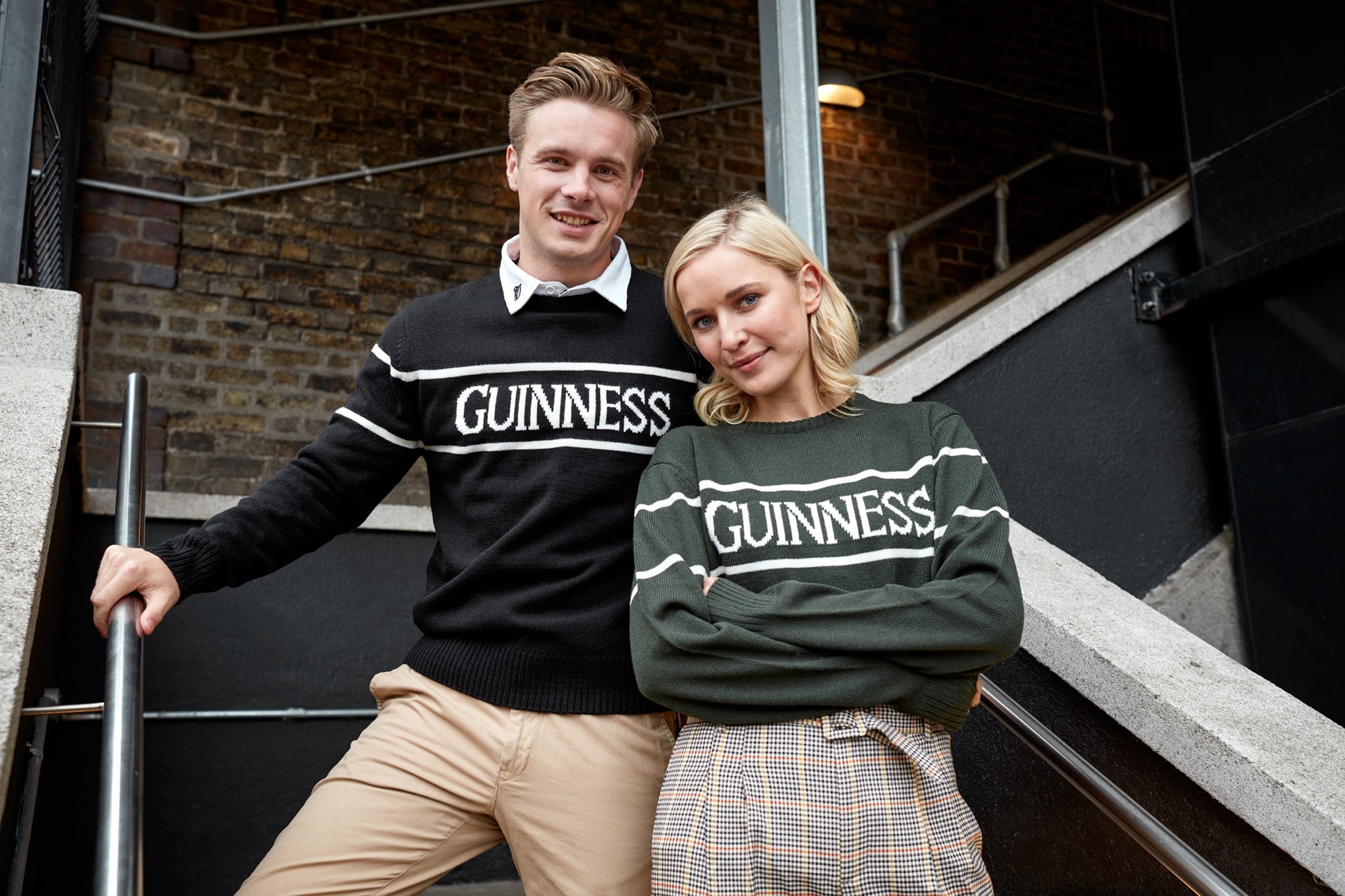 Two people wearing Guinness UK branded Guinness Bottle Green Crew Neck Jumpers posing on stairs with a brick wall background.