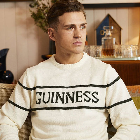 A young man wearing a Guinness Cream Crew Neck Jumper with the word "Guinness" across the chest sits in a warmly lit indoor setting.