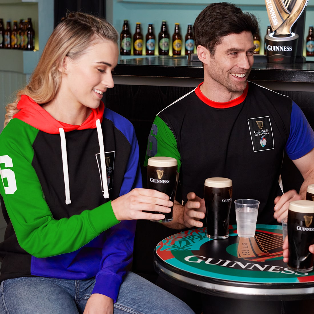 A man and a woman sitting at a table, both wearing Guinness UK Six Nations Colour Block Hoodies in different colors.
