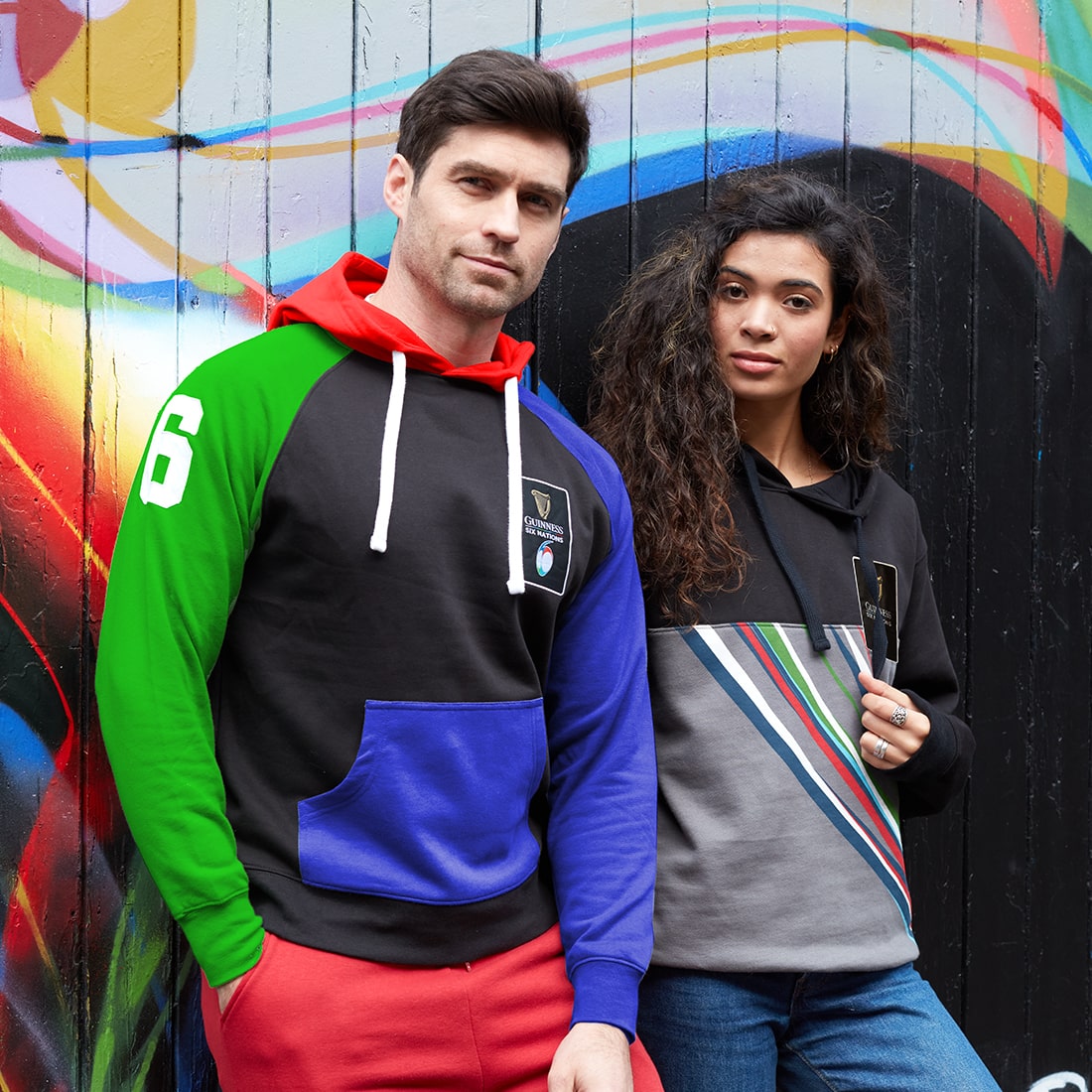 A man and a woman standing next to a graffiti wall, both wearing vibrant Guinness UK Six Nations Colour Block Hoodies.