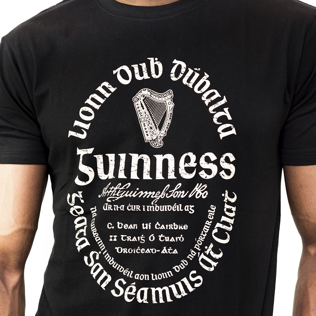 A person wearing the Guinness® Distressed Gaelic Label T-Shirt by Guinness UK, featuring the iconic harp logo and stylized Irish phrases—official Gaelic label-inspired merchandise.