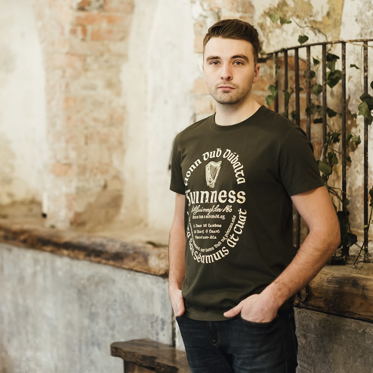 A man stands indoors against a rustic wall, wearing jeans and a Khaki Green Gaelic Label Tee by Guinness UK. This unisex cotton t-shirt features the iconic Trademark Label for a classic look.