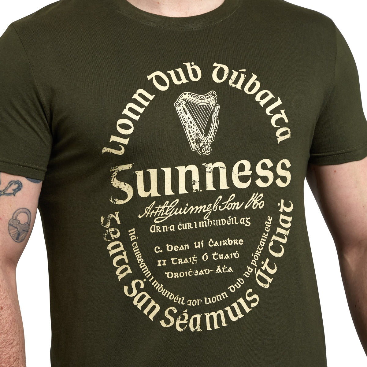 A person wears the Khaki Green Gaelic Label Tee by Guinness UK, a unisex cotton shirt featuring a circular design with classic Irish text, trademark label, and central harp symbol.
