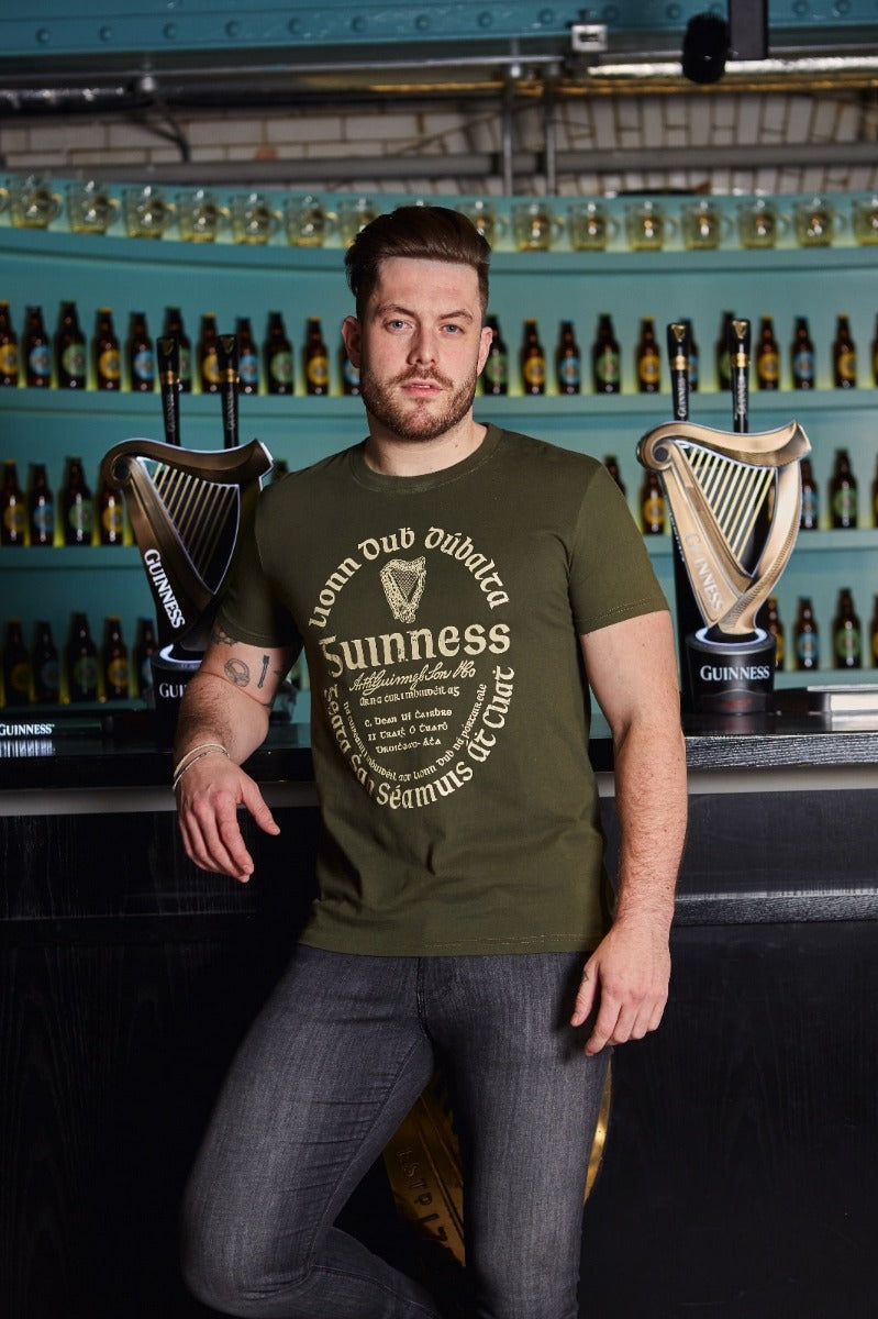 A man with short hair and a beard stands at a bar wearing a Guinness UK Khaki Green Gaelic Label Tee and jeans. Behind him, shelves of beer bottles and Guinness Trademark Label taps are visible.