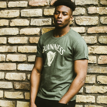 A man wearing a Guinness UK Green Vintage Heathered Harp T-shirt leaning against a brick wall.