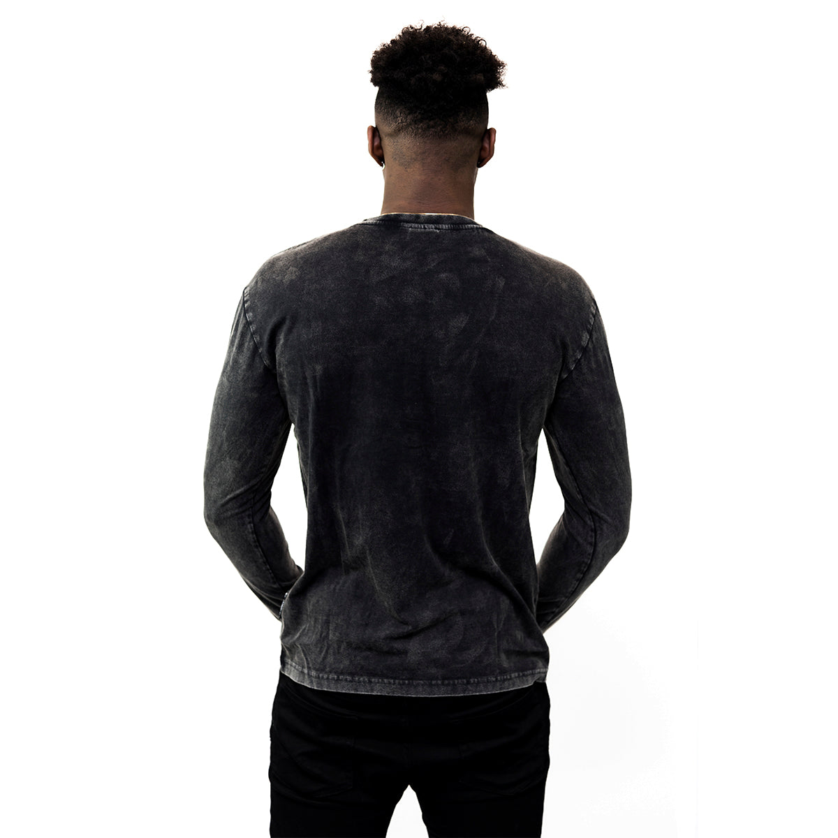 A person with short curly hair stands facing away from the camera, wearing a faded black Guinness Classic Henley and black pants against a white background.