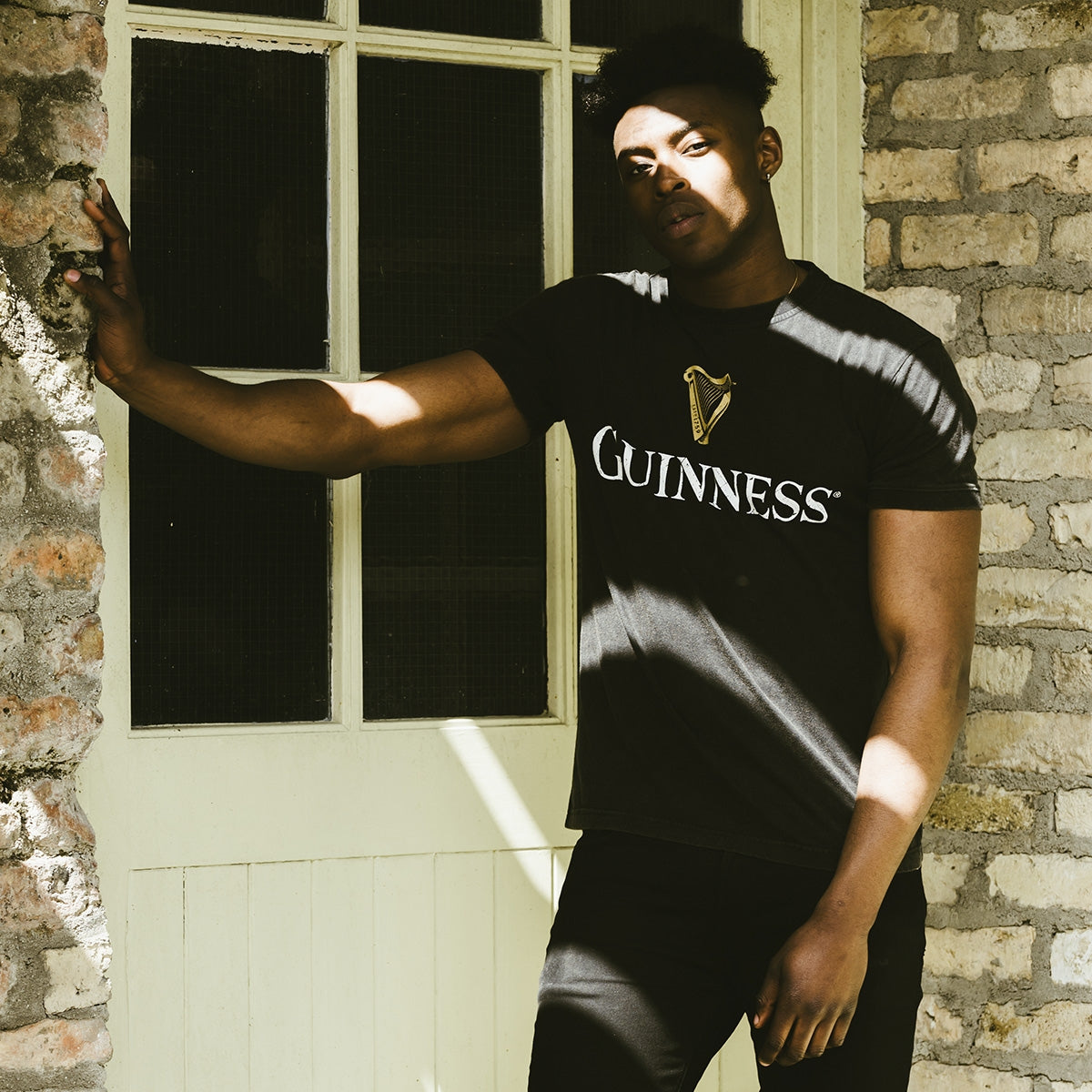 A man leaning against a distressed brick wall wearing a Guinness Distressed Trademark Label T-Shirt.