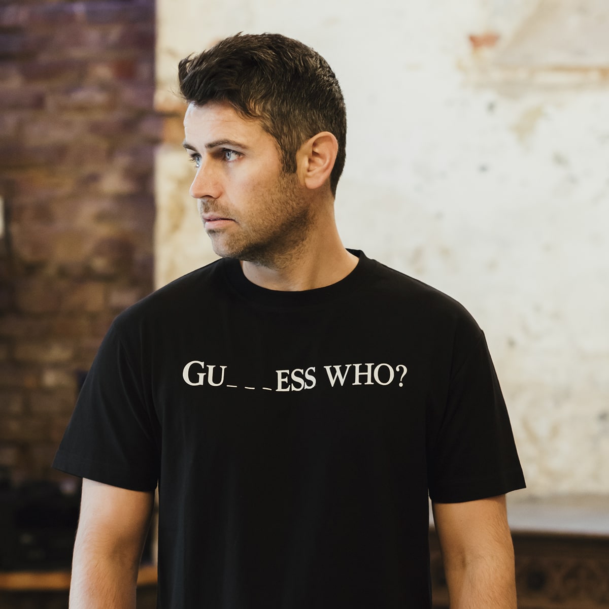A person stands indoors against a blurred brick wall, wearing a black "Guess Who" Tee by Guinness UK. The tee features a distressed finish with the text "GU__ESS WHO?" prominently displayed.