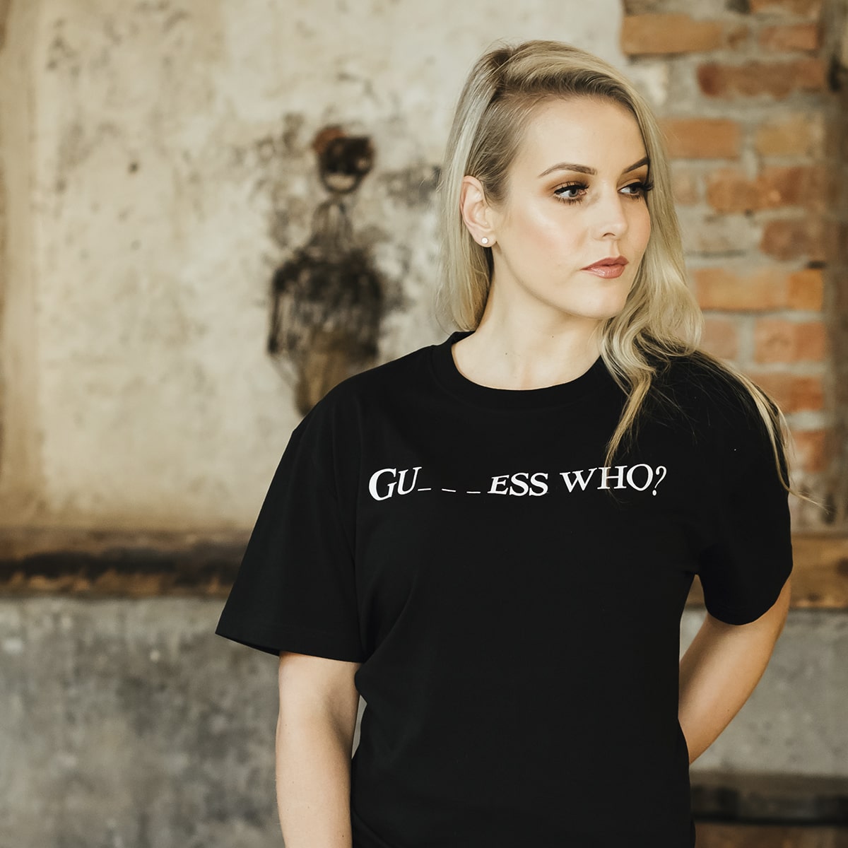 A woman with long blonde hair wears a black "Guess Who" Tee by Guinness UK, standing in front of a weathered brick and plaster wall.