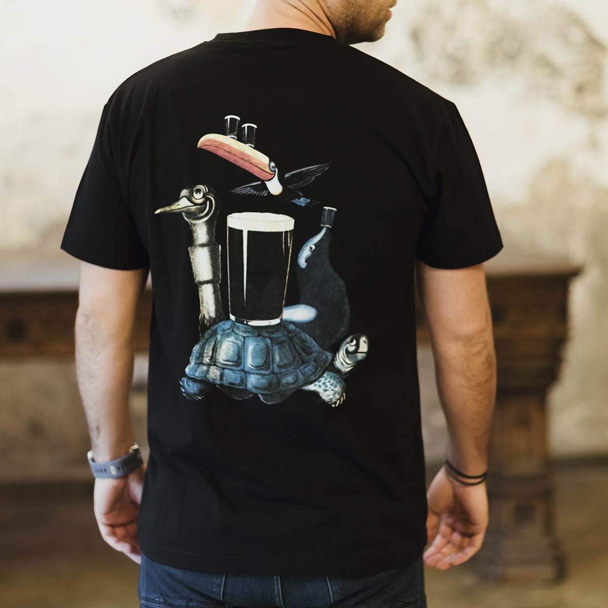 Someone dressed in the "Guess Who" Tee by Guinness UK, featuring a black distressed finish with a design of a bird, a pint of beer, and a turtle on the back, embodies the iconic style reminiscent of Gilroy Guinness illustrators.