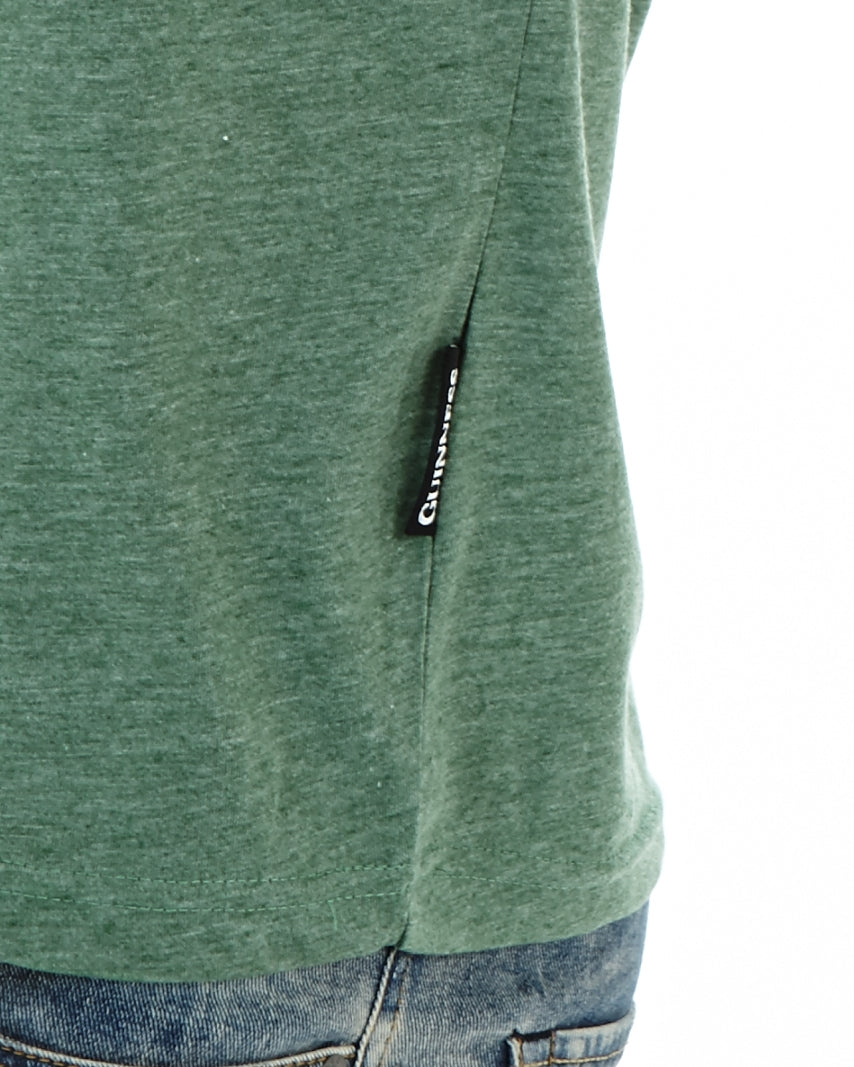 A close-up of a person wearing the Guinness Extra Stout Label Green T-Shirt by Guinness UK, with a visible black tag on the side, paired with blue jeans. The high-quality poly cotton fiber and inclusive size range ensure comfort and style for everyone.