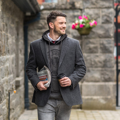 A man with a Guinness hoodie with bottle pocket walking down the street with a coffee in his hand, while his bottle pocket holds a Guinness.