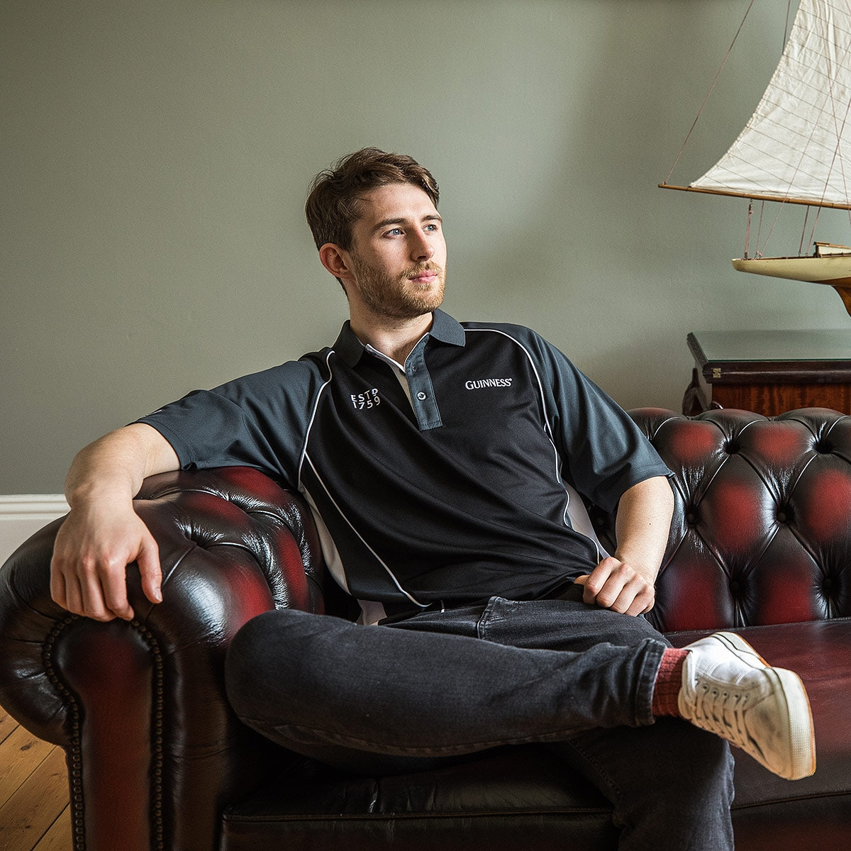 A man in a black Guinness Golf Shirt sits on a dark red leather couch with one arm resting on the backrest. A model sailboat is visible in the background.