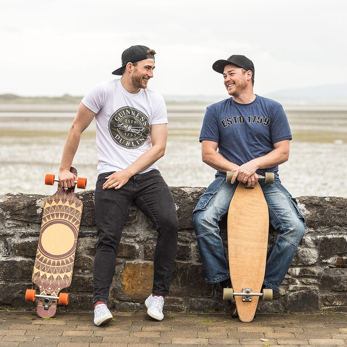 Two men sitting on a wall with their skateboards, wearing Guinness UK's Navy Heathered EST 1759 T-Shirts.
