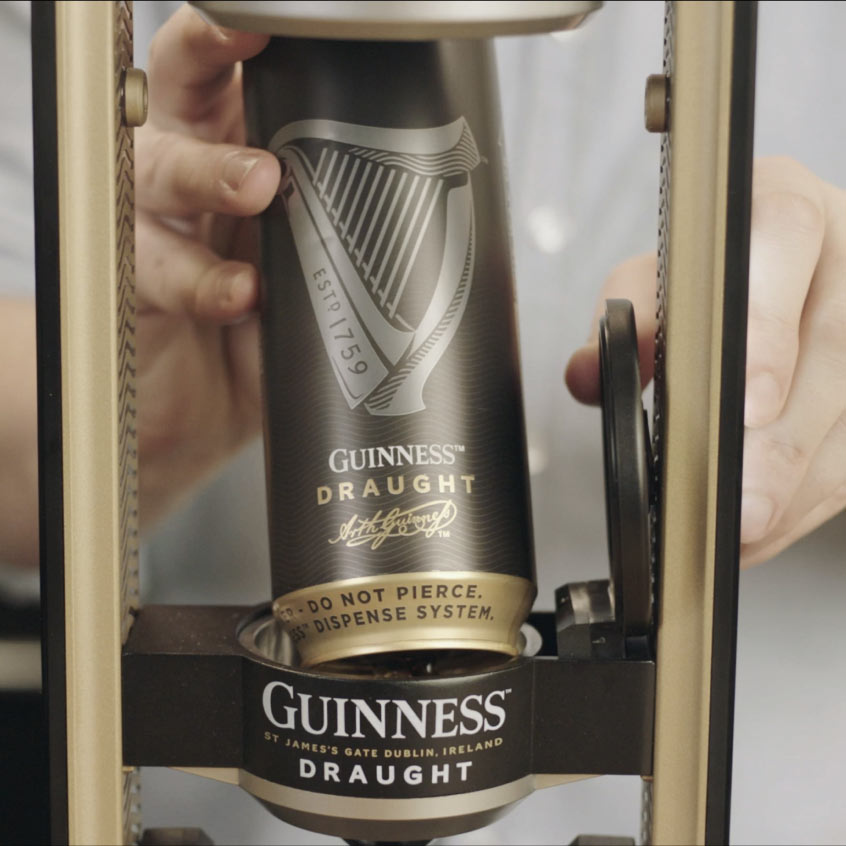 A person placing a can of Guinness Draught into a Guinness UK Guinness MicroDraught device at their home bar. The can, featuring the harp logo and text stating “Estd. 1759” and “Do Not Pierce. Uses Widget Inside For Dispense System,” promises the perfect pint of Draught Guinness®.