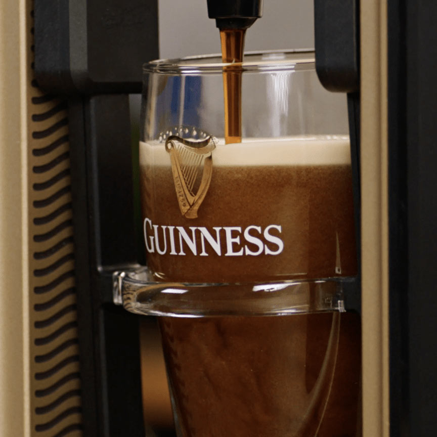 A glass with the Guinness logo is being filled with beer from a tap, creating a dark brew with creamy foam on top—your perfect Draught Guinness® experience, now achievable with the innovative Guinness MicroDraught from Guinness UK for your home bar.