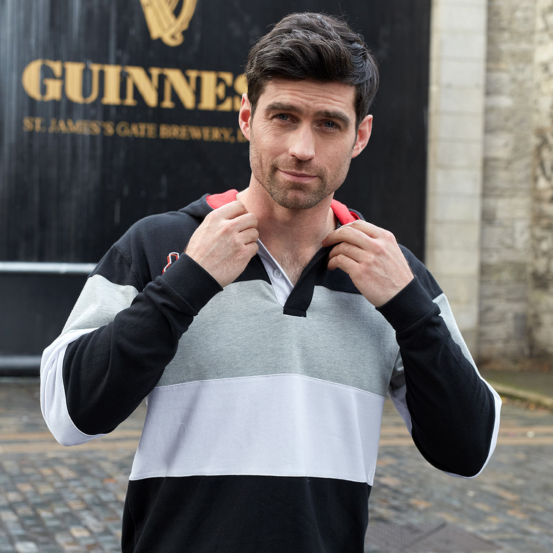 A hooded man is posing in front of a Guinness UK Black & Red Toucan Hooded Rugby sign.