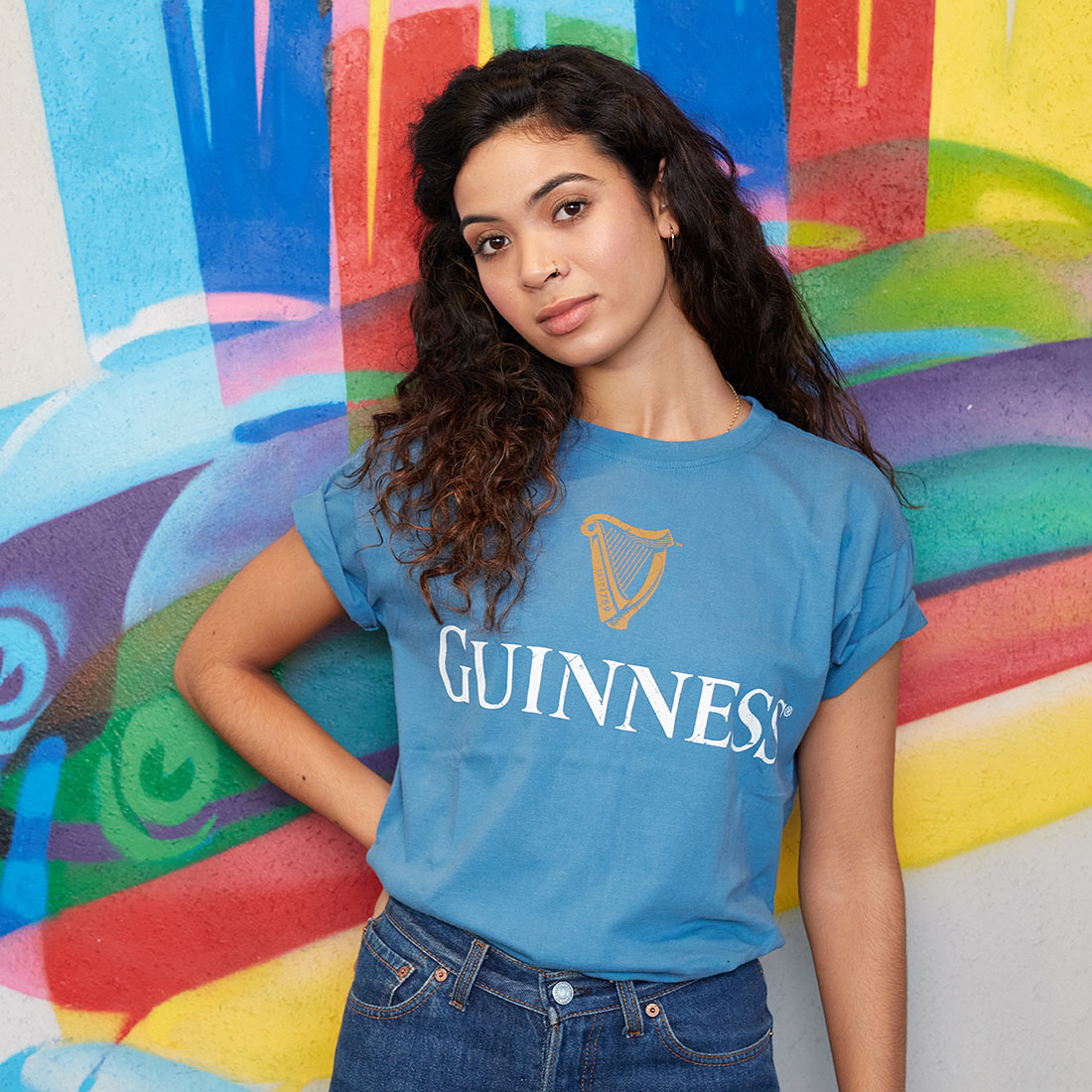 A woman sporting a Sky Blue Guinness Harp Premium T-Shirt poses confidently in front of a vibrant, colorful wall, representing the brand Guinness UK.