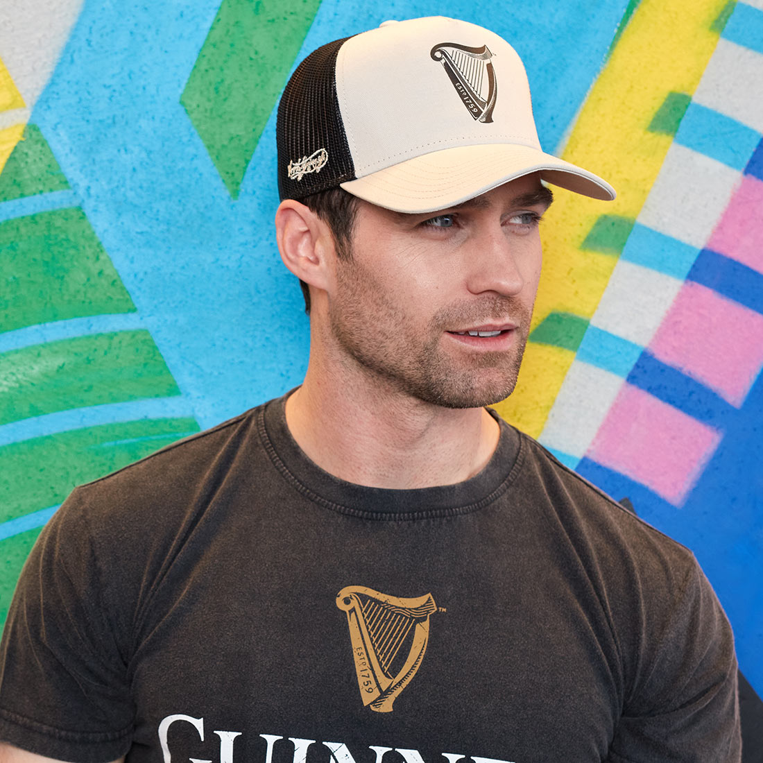 A man wearing a Guinness UK hat in front of a colorful wall decorated with the Guinness UK logo, featuring the Guinness Premium Beige with Black Harp Cap.