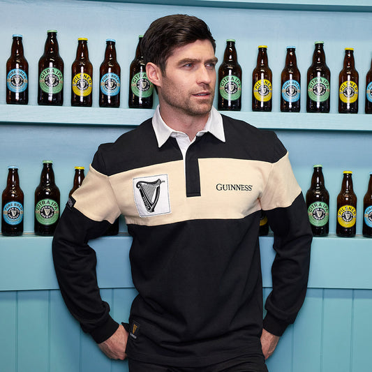 A man wearing a Guinness traditional rugby jersey with cream panel and harp logo patch is leaning against a wall of Guinness beer bottles.