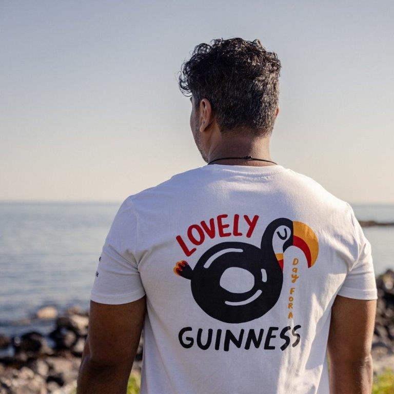 A man wearing a FATTI BURKE "LOVELY DAY FOR A GUINNESS" TOUCAN WHITE TEE featuring the renowned Guinness logo at a lively summer festival.