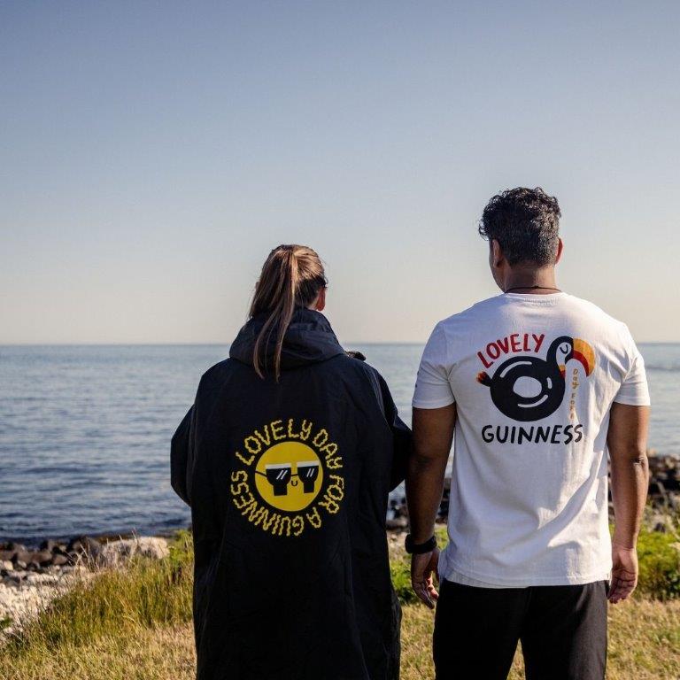 Two people standing next to the ocean wearing FATTI BURKE "LOVELY DAY FOR A GUINNESS" TOUCAN WHITE TEE from Guinness Webstore UK.