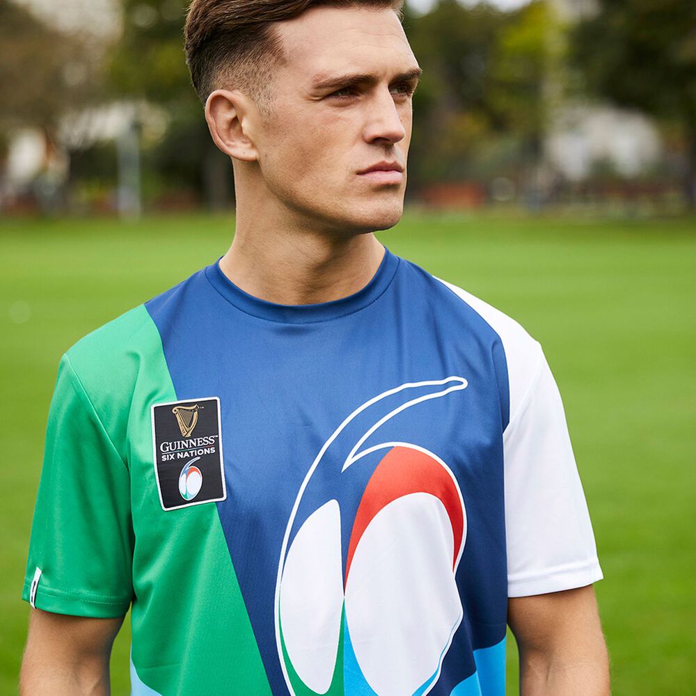 A person stands on a grassy field, wearing a Guinness Webstore UK Multi-coloured Performance T-shirt with a Guinness Six Nations logo. Trees and a blurred background are visible.