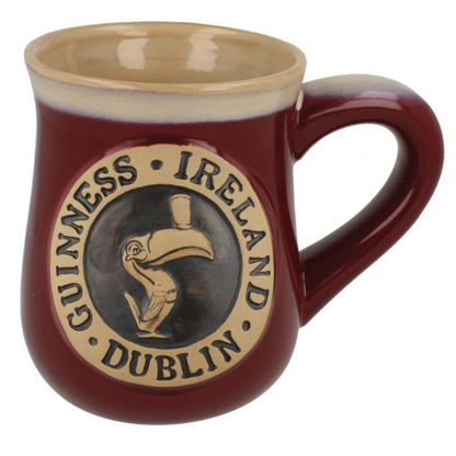 A Guinness Toucan Burgundy Pottery Mug with a handle features "Guinness Ireland Dublin" and a toucan logo on a beige medallion set against a maroon background.