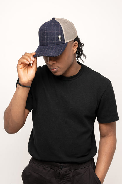 A person in a black t-shirt and dark pants tilts the brim of a Guinness Checked Trucker Cap by Guinness Webstore UK, set against a plain light background.