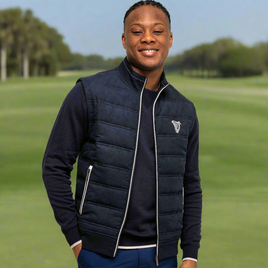 A man smiles on a golf course with his hands in his pockets, wearing the Guinness Performance Gilet from the Guinness Webstore UK, featuring embroidered branding.