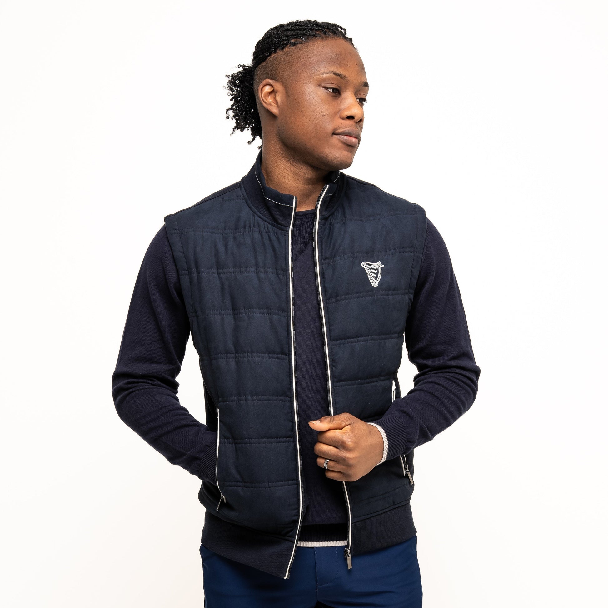 A person models the navy Guinness Performance Gilet from Guinness Webstore UK, featuring quilted detailing, over a dark long-sleeve shirt and blue shorts against a plain white background.