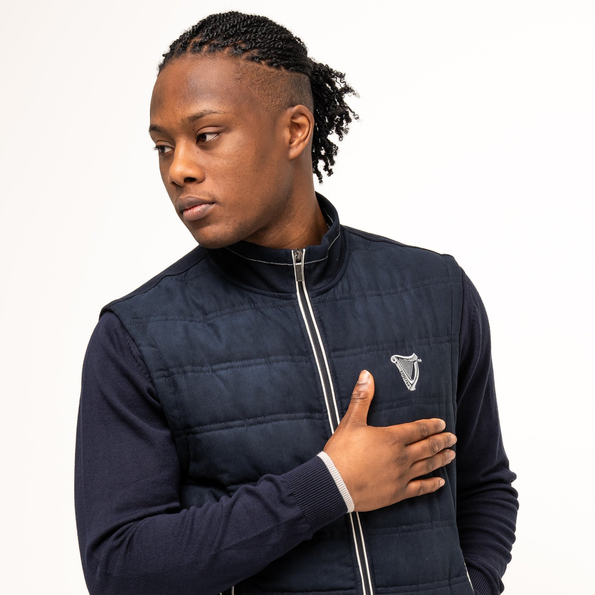 A man stands against a plain background, wearing the Guinness Performance Gilet from Guinness Webstore UK over a dark sweater, with one hand on his chest and looking to the side.