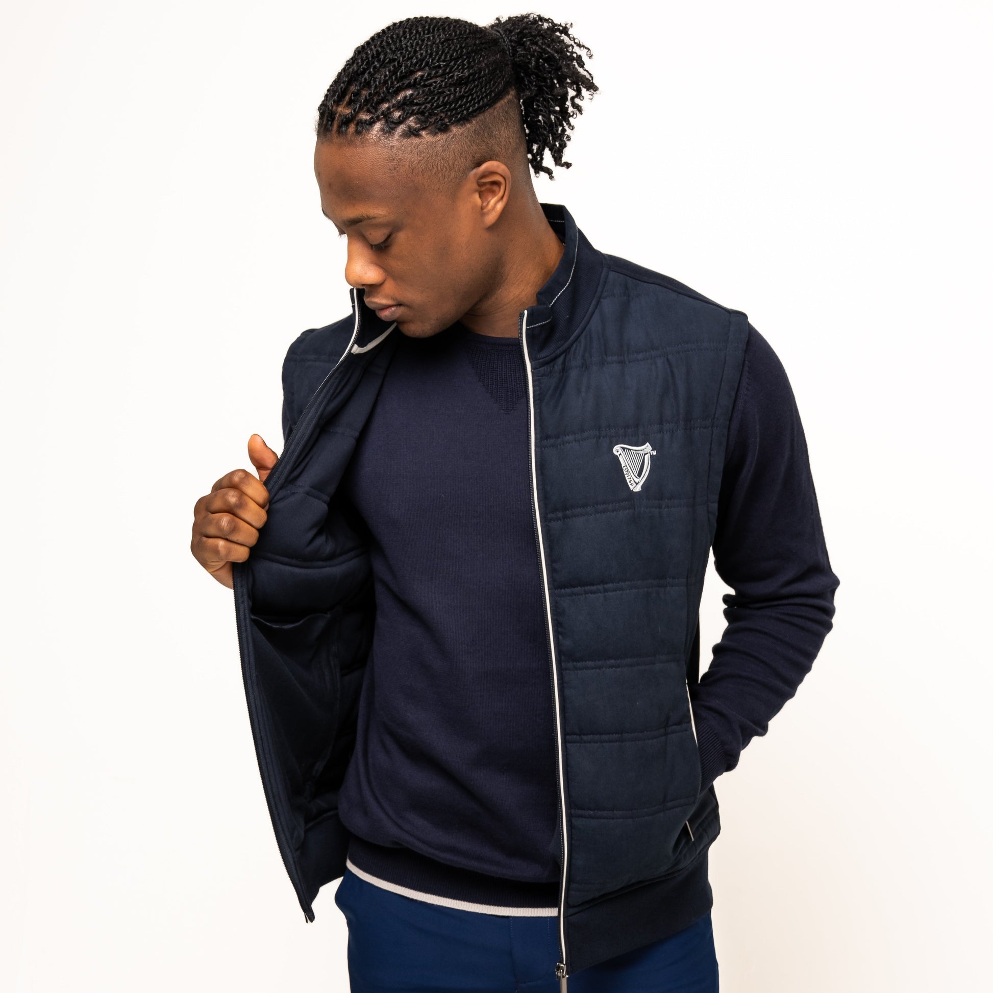 A man wears the Guinness Performance Gilet from Guinness Webstore UK, featuring quilted detailing, over a navy sweatshirt and blue shorts. He stands against a white background, looking down as he opens the gilet.