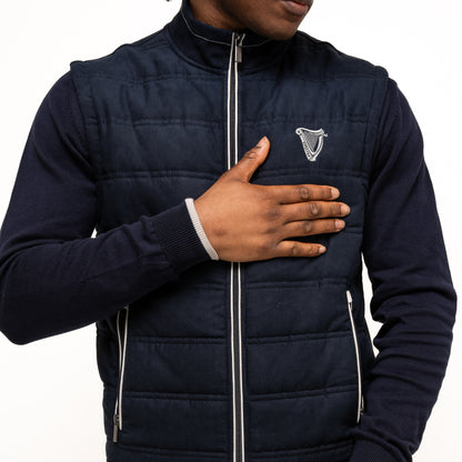 A person wearing the navy Guinness Performance Gilet from Guinness Webstore UK, featuring quilted detailing, a navy sweater, and blue pants places one hand on their chest and the other by their waist.
