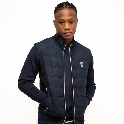 A man wearing a Guinness Webstore UK Performance Gilet with quilted detailing over a dark long-sleeve shirt and blue shorts stands with his hands in his pockets against a white background.