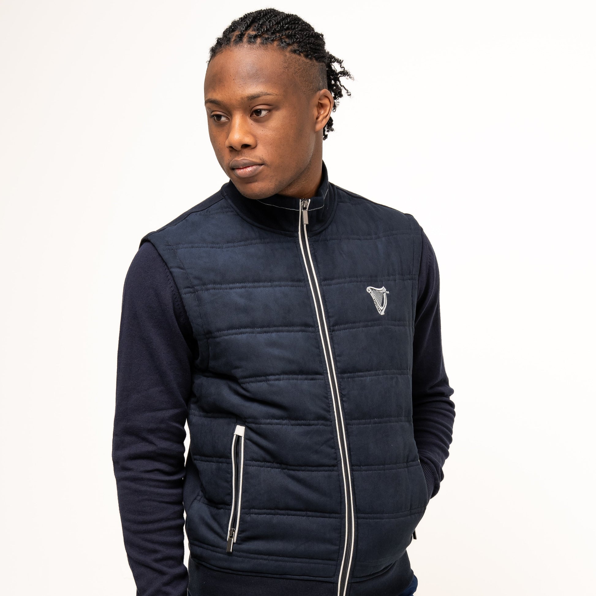 A man models the navy Guinness Performance Gilet from Guinness Webstore UK, featuring quilted detailing, over a dark zip-up sweater and blue shorts, standing against a plain white background and looking to his left.