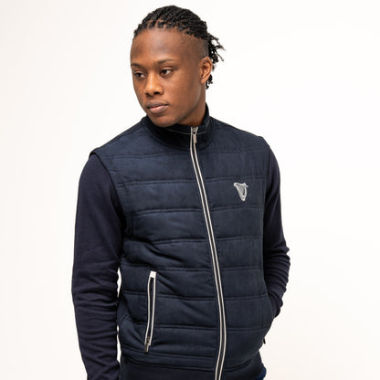 A man models the navy Guinness Performance Gilet from Guinness Webstore UK, featuring quilted detailing, over a dark zip-up sweater and blue shorts, standing against a plain white background and looking to his left.