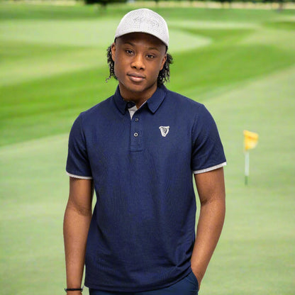 Wearing the Guinness Performance Polo - Navy by Guinness Webstore UK, along with navy shorts and a light gray cap, a person stands against a plain white background, one hand in their pocket.