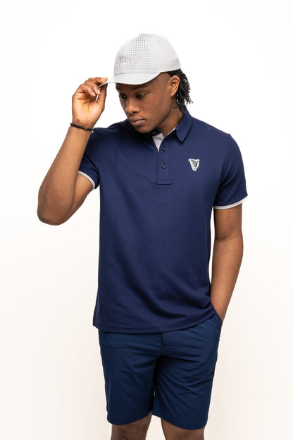 A man wearing the Guinness Performance Polo - Navy from Guinness Webstore UK, navy shorts, and a light gray cap stands against a plain white background, holding the brim of his cap with his right hand.