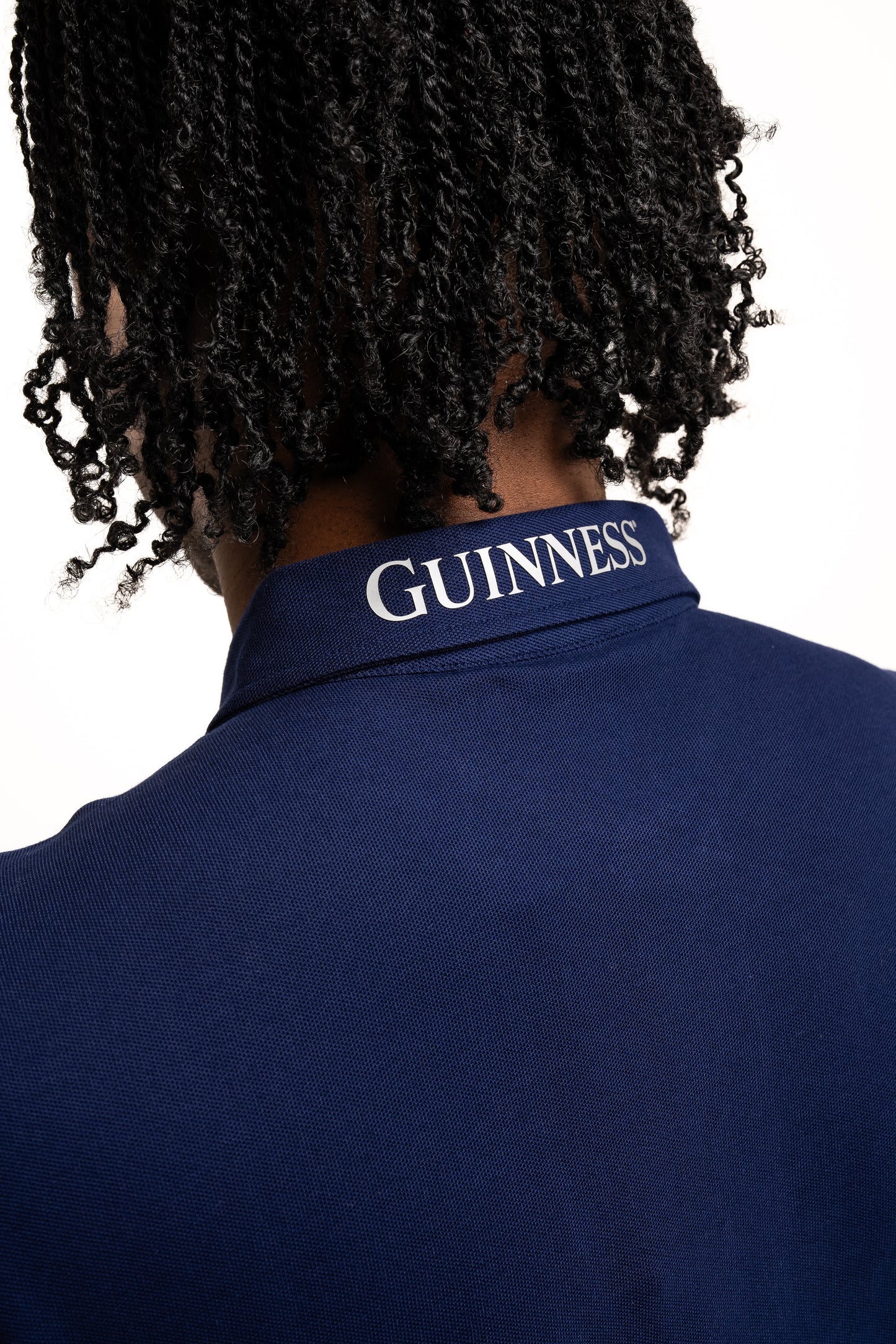 A person with braided hair wears the Guinness Performance Polo - Navy from Guinness Webstore UK, made from premium 210gsm double pique fabric and featuring "GUINNESS" printed on the collar.