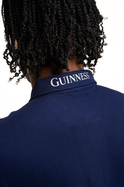 A person with braided hair wears the Guinness Performance Polo - Navy from Guinness Webstore UK, made from premium 210gsm double pique fabric and featuring "GUINNESS" printed on the collar.