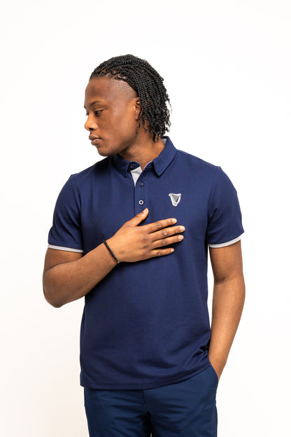 A man wears the Guinness Performance Polo - Navy from Guinness Webstore UK, featuring a crest logo. Crafted with premium double pique fabric, this polo offers all-day comfort. He stands against a white background, looking to his left.