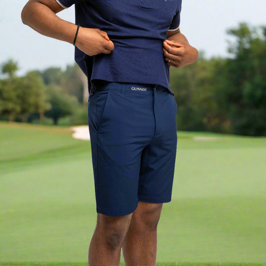 On a golf course with grass and trees in the background, a person wearing a navy blue shirt and Guinness Webstore UK's Guinness Performance Shorts - Navy with a stretch waistband holds up their shirt slightly.