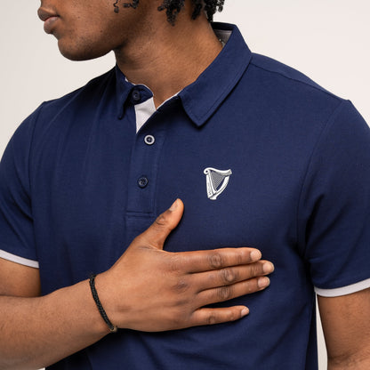A person wearing the Guinness Performance Polo - Navy from Guinness Webstore UK, featuring an embroidered harp logo and crafted from 210gsm double pique fabric, holds their right hand over the left side of their chest.